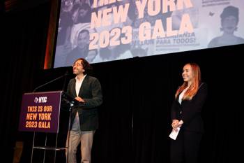 Two people smile from a stage with New York Immigration Coalition 2023 gala signage in the background and on a podium.